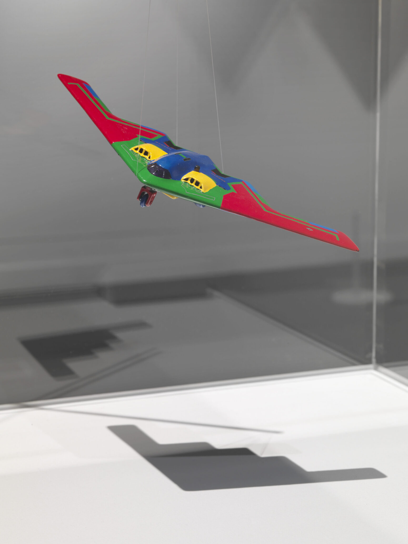 A photo of a colorful red, green, blue, and yellow plane, suspended by some fish line and casting a shadow on the floor.
