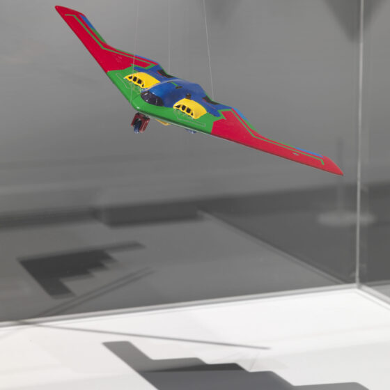 A photo of a colorful red, green, blue, and yellow plane, suspended by some fish line and casting a shadow on the floor.