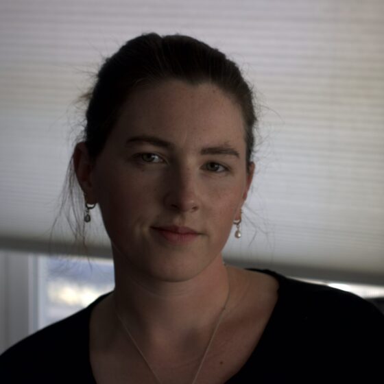 A Palestinian-American woman with straight, brown hair pulled back and dark eyes. She stands in a dimly lit room, wearing a black shirt.