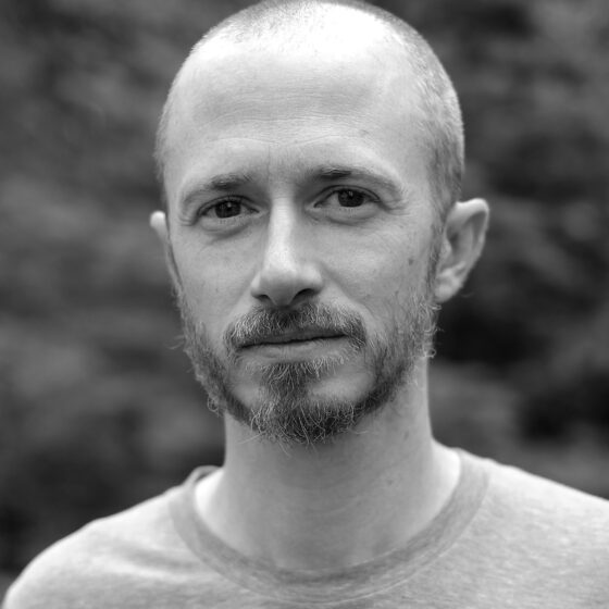 A black-and-white photo bald man with a short beard. He stands outside and wears a light-colored T-shirt