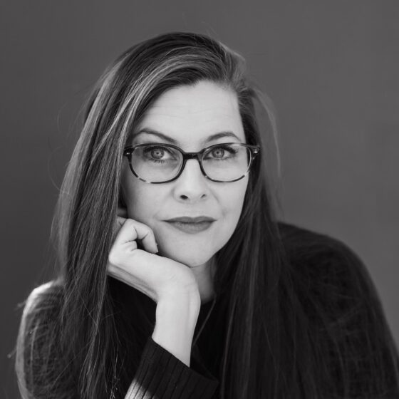 A black-and-white photo of a White woman with long, straight, dark hair and dark eyes. She rests her chin on her right hand, wearing a black, long-sleeved shirt and cat-eye glasses.