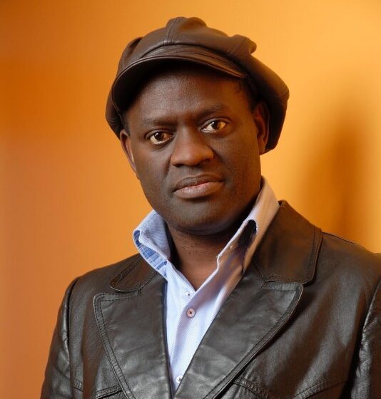 A man with brown eyes standing in front of an orange wall and wearing a black, leather jacket, a white, collared shirt, and a brown, floppy hat