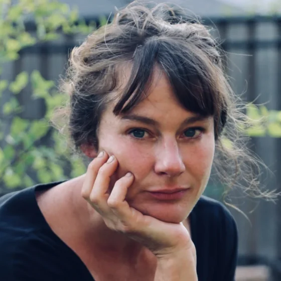 A woman with straight, brown hair tied up in a bun and blue eyes. She sits outside, holding her chin in her right hand, and wearing a black shirt