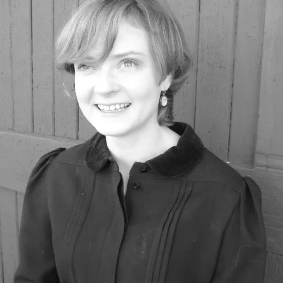 A black-and-white photo of a woman with short, straight, light-colored hair, smiling. She stands in front of a wooden fence and wears a dark-colored, collared shirt.