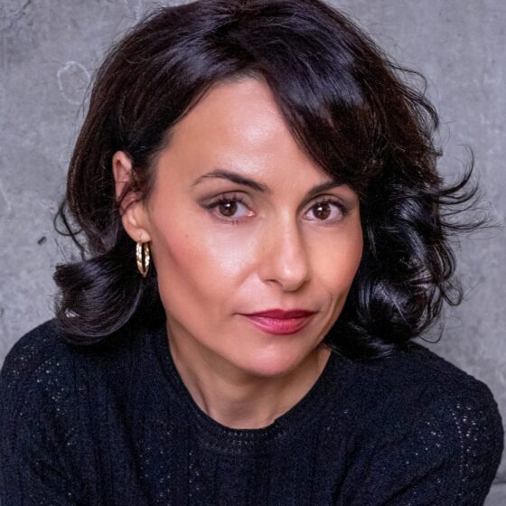 A woman with short, black, curly hair and dark-brown eyes. She sits in front of a gray background and wears a black shirt