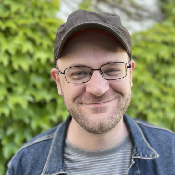 A man with a short, brown beard and blue eyes, smiling. He stands outside in front of a bush, wearing a gray-striped shirt, a jean jacket, a gray hat, and rectangular glasses