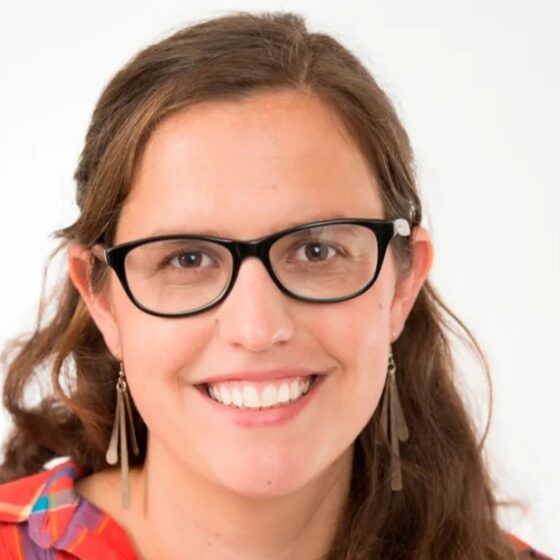 A woman with long, straight, dirty-blonde hair and brown eyes, smiling. She stands in front of a white background and wears black, cat-eye glasses and a red plaid shirt