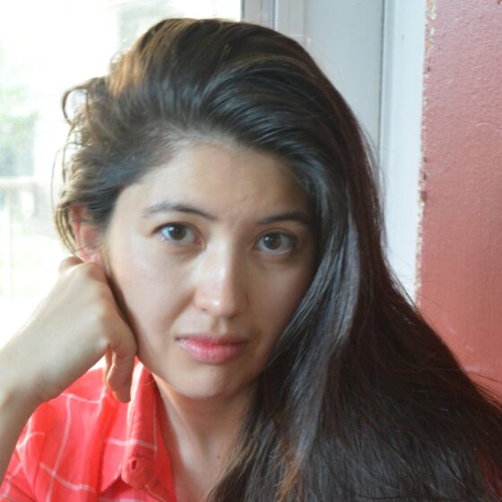 An Asian-American woman with long, straight, black hair and brown eyes. She rests her cheek on her right hand, and wears a coral, collared shirt