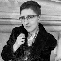 A black-and-white photo of a woman with short, dark hair and dark eyes, smiling. She wears a printed shirt, a dark cardigan, and cat-eye glasses