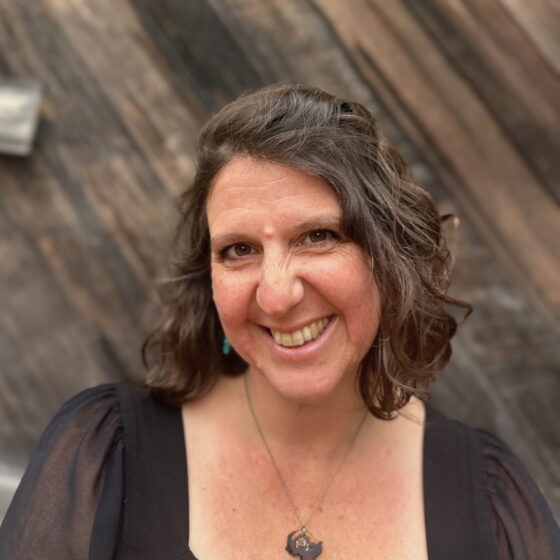 Maya Jewell Zeller, a white woman with short brown hair, wears a black square-necked top and pendant necklace. She smiles broadly in front of an aged wood background.