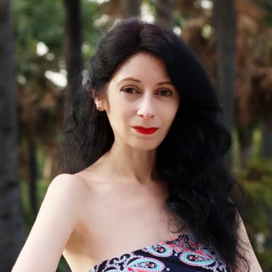 A Hispanic woman with long, fluffy, black hair worn half-back and brown eyes. She stands outside, wearing a black, strapless dress with a pink-and-blue pattern.
