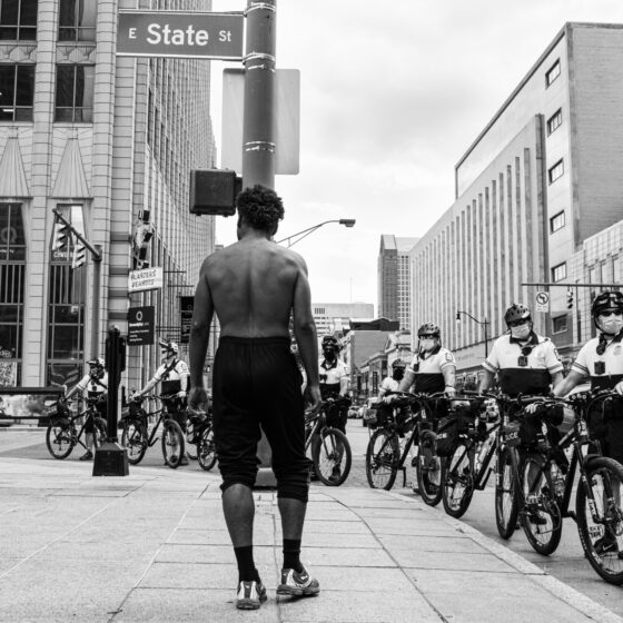 A black-and-white photo of a shirtless man on a city sidewalk, facing a line of police-bikers