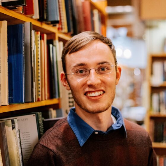 A man with short, straight, blonde hair and brown eyes, smiling. He stands in a library, wearing a brown sweater with a blue, collared shirt.
