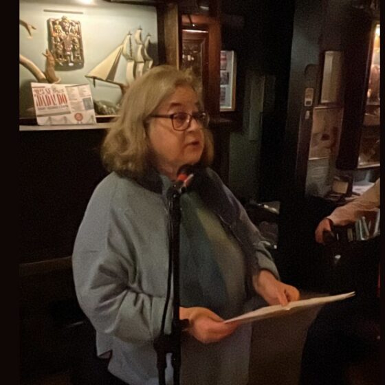 Carol Dorf stands at a lectern wearing spectacles and a sweater