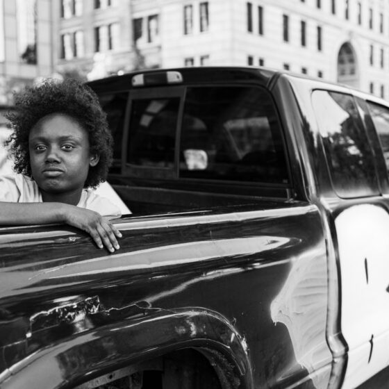 A black-and-white photo of a young girl in the bed of a black pickup truck with "BLM" spray-painted on the side