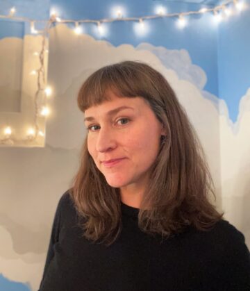 A white woman with light brown hair that reaches her shoulders. She's wearing a dark shirt and standing in front of a mural of clouds and sky, with small lights on a string.