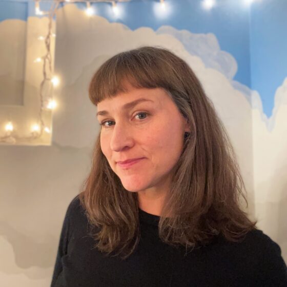A white woman with light brown hair that reaches her shoulders. She's wearing a dark shirt and standing in front of a mural of clouds and sky, with small lights on a string.