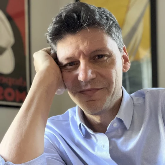 A man with short, curly, salt-and-pepper hair and dark eyes, smiling. He leans his head against his right hand and wears a light-blue collared shirt.