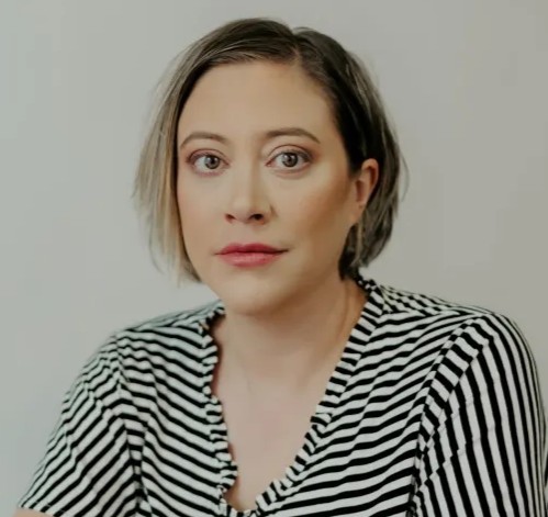 A woman with short, straight hair that fades from brown to blonde. She has brown eyes and sits in front of a white background. She wears a black-and-white-striped shirt.