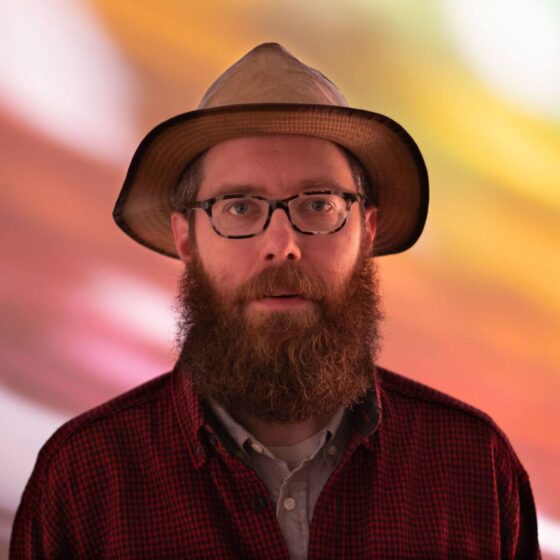 A man with a long, red beard and blue eyes. He stands in front of a rainbow-like background, wearing a collared shirt, a brown, corduroy jacket, and a brown, trilby hat