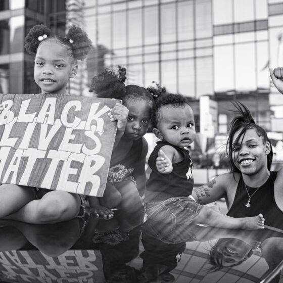 A black-and-white photo of a little girl holding a sign that says "Black Lives Matter," sitting on a car with two other small children and a woman standing next to them, smiling and holding her fist in the air