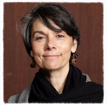 A woman with short, straight, salt-and-pepper hair and brown eyes, smiling. She wears a gray sweater and a black scarf in front of a brown background