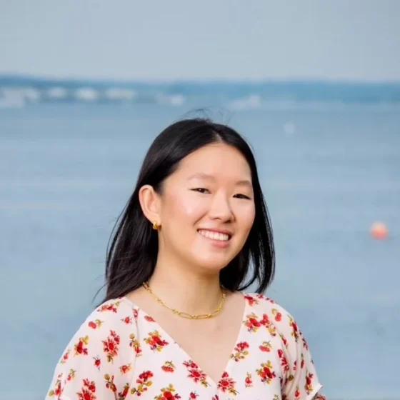 A woman with straight, shoulder-length, black hair, smiling. She stands in front of a beach, wearing a pink shirt with red flowers on it