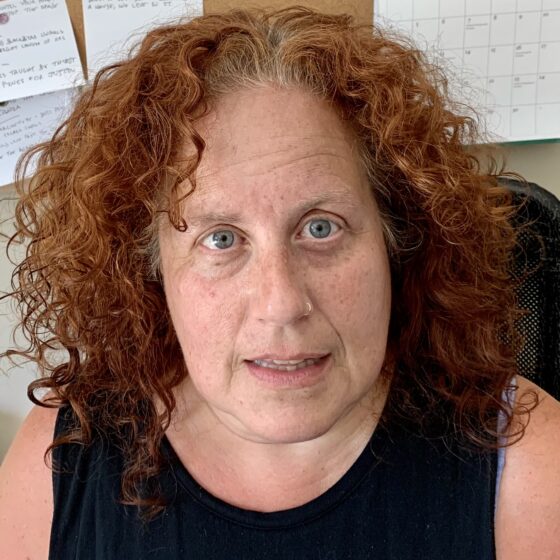 A white woman with curly red hair and blue eyes. She's wearing a dark sleeveless shirt and is front of a bulletin board with a few items tacked to it