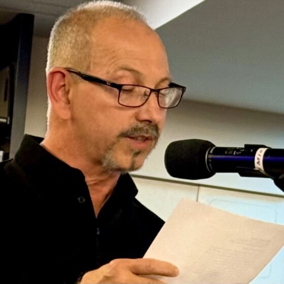 A White man with buzzed, gray hair and a goatee. He wears a black, polo shirt and rectangular glasses. He is reading off of a paper into a microphone.