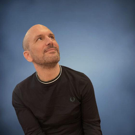 A man with stubble smiling and gazing upward, to the side. He is wearing a black long-sleeved tee and white pants, and seated against a blue backdrop.