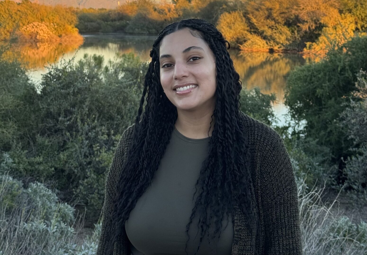 A Black woman smiling at the camera. She is standing by foliage and a body of water, and is wearing a gray cardigan.