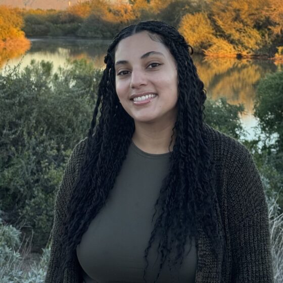 A Black woman smiling at the camera. She is standing by foliage and a body of water, and is wearing a gray cardigan.