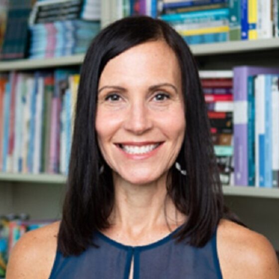 A woman with straight, dark, shoulder-length hair and brown eyes, smiling. She stands in front of a library bookshelf and wears a sheer, blue tank top