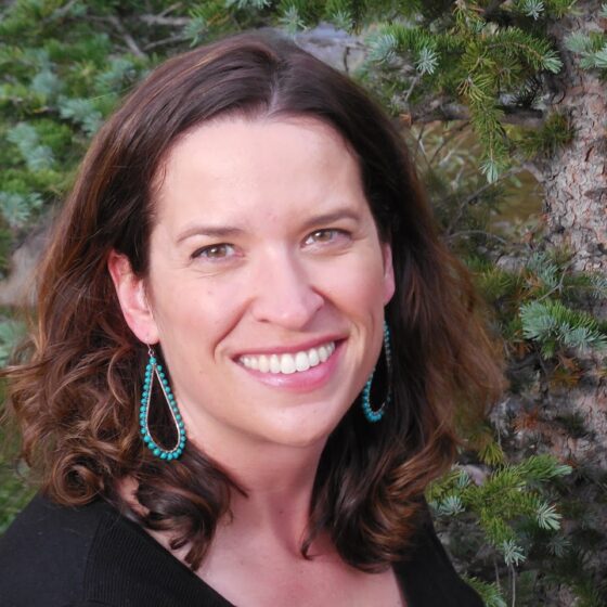 A woman with shoulder-length, straight, brown hair and hazel eyes, smiling. She stands in front of a forest, wearing a black shirt and large, turquoise earrings