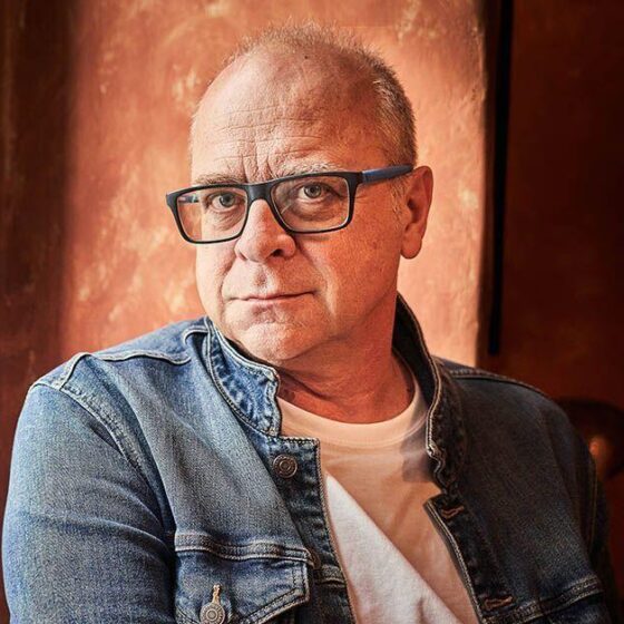 A bald man, smiling. He sits in a room with an orange wall and wears a white shirt, a jean jacket, and large, square glasses