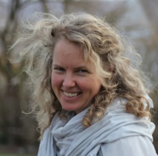 A woman with long, blonde, curly hair blowing in the wind. She is smiling and wearing a light-gray scarf