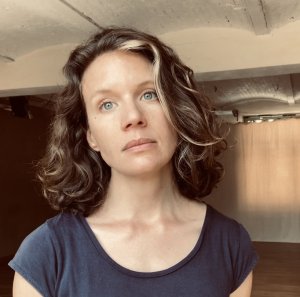 A woman with brown, shoulder-length, wavy hair and blue eyes. She stands in a studio wearing a navy T-shirt.