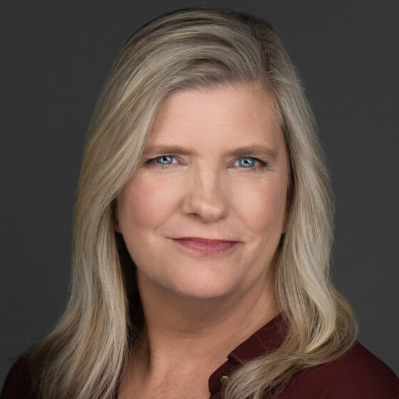 Headshot of a white woman with blond hair and blue eyes. She's wearing a maroon collared and buttoned shirt and is in front of a dark gray background