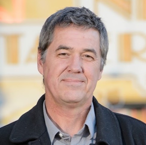 A man with gray hair and brown eyes, smiling. He stands outside of a building, wearing a gray, collared shirt and a black blazer