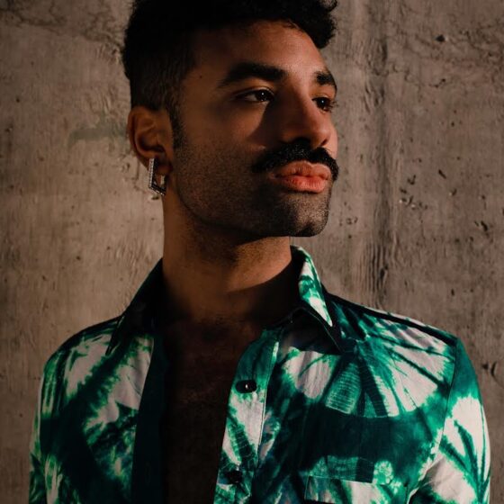 A man with short, dark, curly hair and a mustache, smiling. He wears a black, collared shirt underneath a green-and-white-dyed jacket