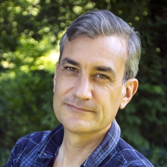 A White man with short, gray hair and brown eyes. He stands outside, wearing a blue-and-black, plaid shirt.