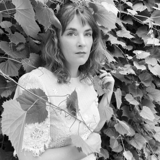 A black-and-white photo of a woman with shooulder-length, light-colored, wavy hair and dark eyes. She stands in front of a wall of ivy and wears a light-colored, lace shirt.