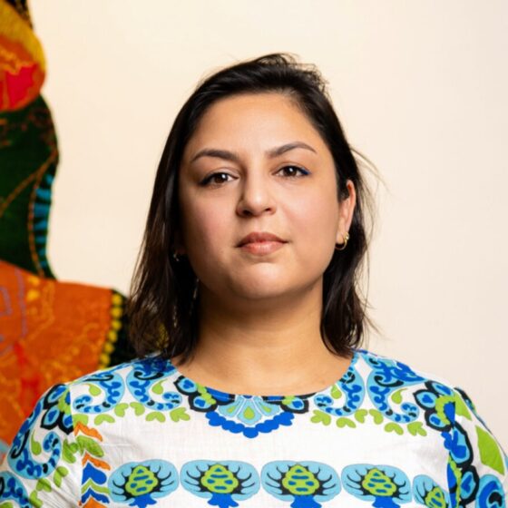 A woman with long, straight, dark-brown hair and brown eyes. She wears a white, blue, and green patterned shirt