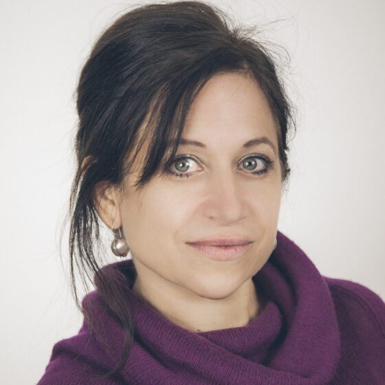 A woman with straight, black hair in an updo and green eyes. She stands in front of a white background, and wears a purple sweater.