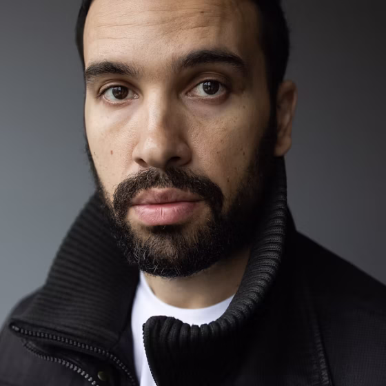 A man with buzzed, black hair, a short, black beard, and brown eyes. He wears a white shirt underneath a black jacket