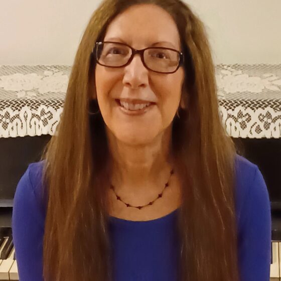 A woman with long, straight, brown hair and brown eyes, smiling. She wears a blue shirt and rectangular glasses