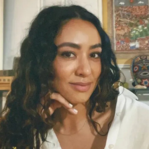 A woman with long, dark, curly hair and brown eyes, smiling. She wears a white, collared shirt