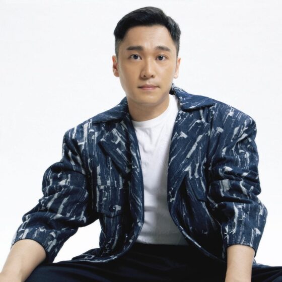 A man with short, straight, black hair and brown eyes. He sits in front of a white background, wearing a patterned, blue-and-black jacket, a white undershirt, and black jeans