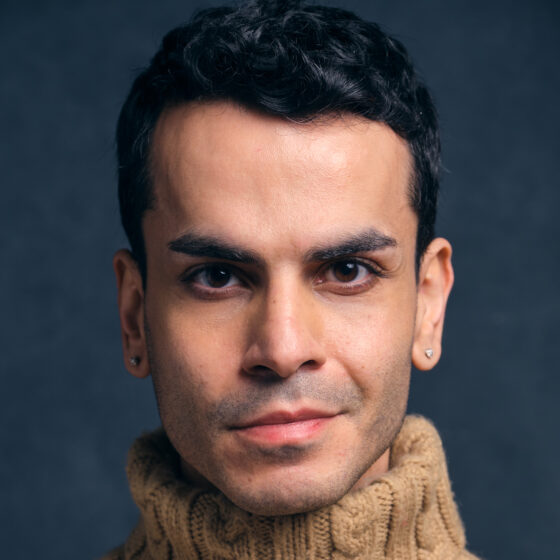 A Middle Eastern/North African person with short dark hair. They're wearing a big knit beige turtleneck and in front of a dark gray background