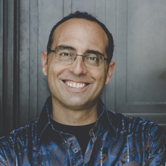 A man with buzzed, black hair and brown eyes, smiling. He wears a black-and-blue, collared jacket and rimless, rectangular glasses